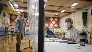 A poll worker behind a sheet of plastic in August
