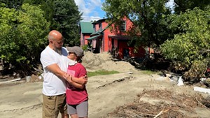 A man and a woman hold each other outside a flood damaged house. They are surrounded by dirt and a washed out yard.