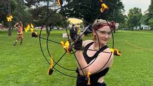 A woman holds a flaming parasol prop.